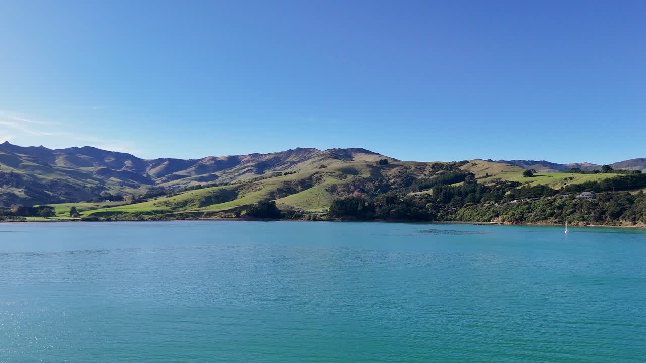 Aerial footage captures Akaroa's serene lake and rolling hills under clear blue skies, showcasing natural beauty and tranquility
