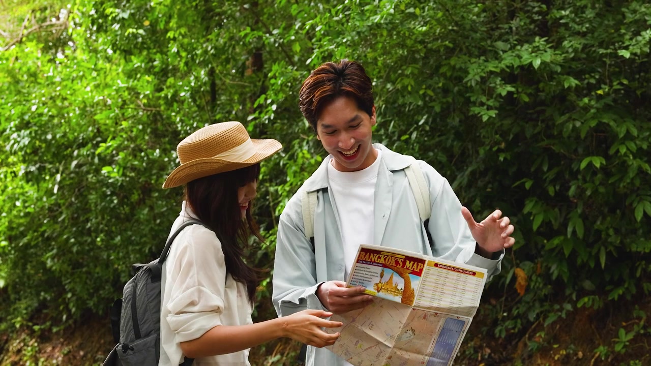 Happy young Asian couple reading a paper map together while hiking along a lush trail.