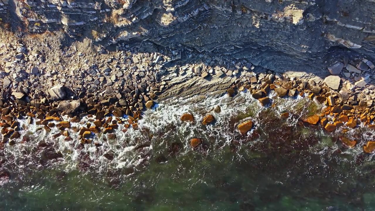 Waves crash against rocks along a coastline with clear water and rocky formations seen from above near the shore at noon