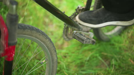 close-up of individual moving bike in grassy field, wearing black trousers, adjusting foot on pedal, lowering leg back onto grass, with focus on pedal and tire in blurred green natural background