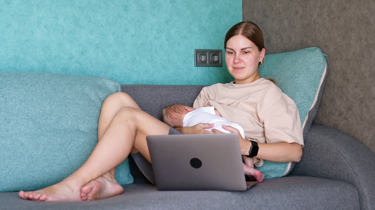 Caucasian young woman combining work and childcare. Mother of infant feeds a baby and works on laptop.
