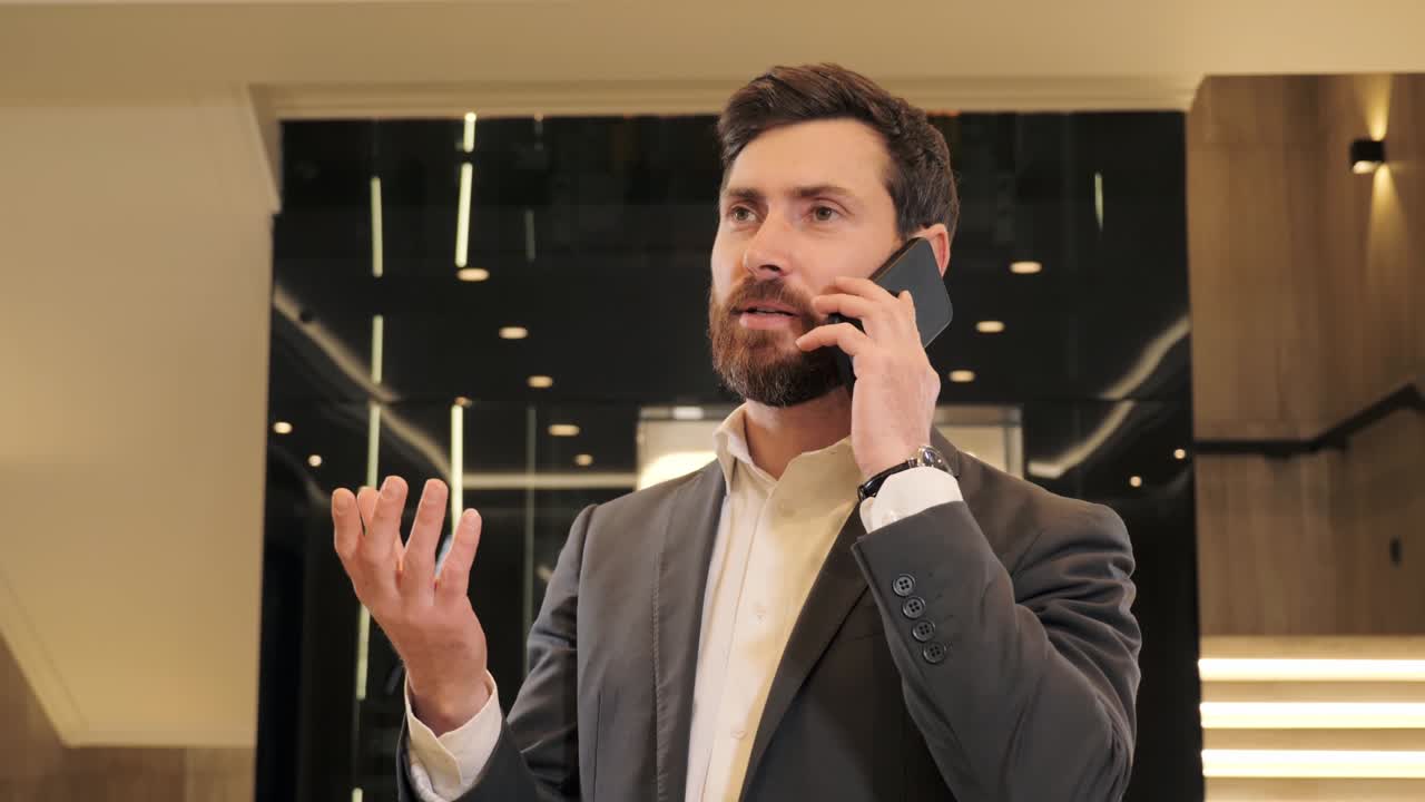 Portrait handsome man having phone call in business center. Businessman talking on smartphone in hall near modern glass elevator. Phone call phone indoor. Office employee using modern device.