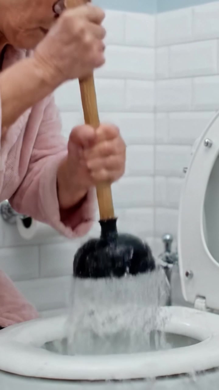 A determined older woman using a plunger to unclog a toilet, showcasing her resolve and expertise in handling household challenges effectively.