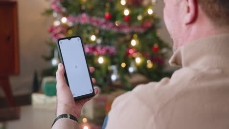 senior man using smartphone by christmas tree, enjoying festive holiday season