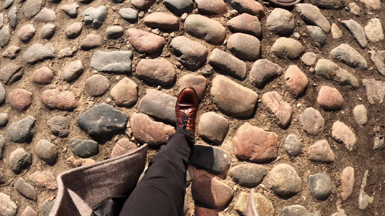 First person view of a businessman walking on a cobblestone pavement wearing elegant brown shoes and a beige coat, with a manhole cover in the background in slow motion