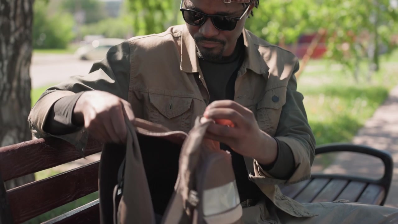 Close up of man sitting on bench in park adjusting headphones and opening bag. Man interacts with personal items while seated outdoors surrounded by greenery and soft daylight. Background