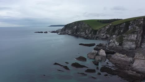 Aerial-Drone-Shot-of-Ayrmer-Cove-in-Devon,-England-with-calm-crystal-blue-waters-and-stunning-cliffs-,-rolling-green-countryside-and-seagulls-in-shot