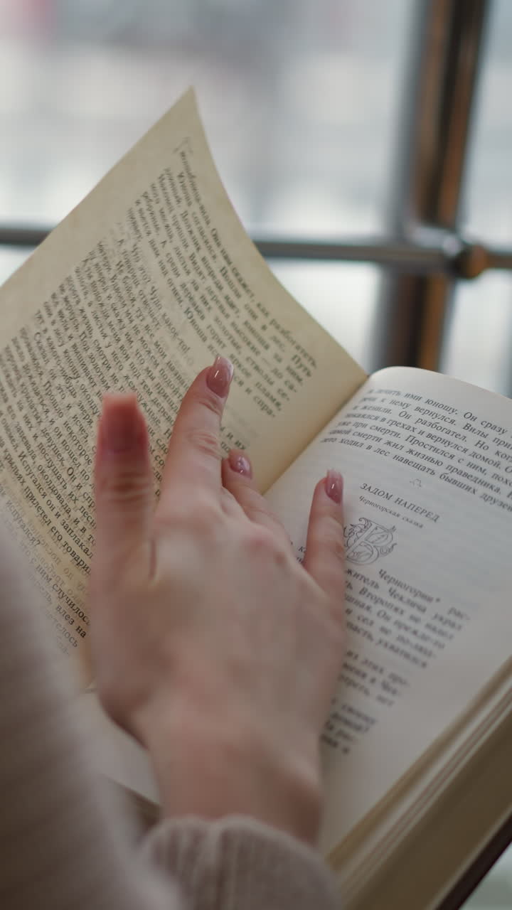 vista trasera de primer plano de la dama volviendo a una nueva página en el libro con una taza de té en la parte delantera, borroso asentamiento urbano ocupado visible en el fondo a través de la ventana