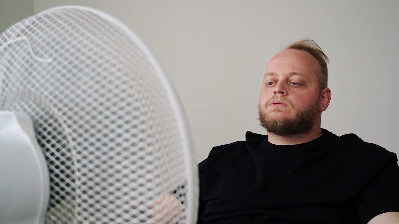 Uomo sudato in maglietta nera si accascia davanti a un ventilatore che gira, cercando di rinfrescarsi in una stanza calda. Concetto per ondata di calore, spossatezza e raffreddamento domestico.