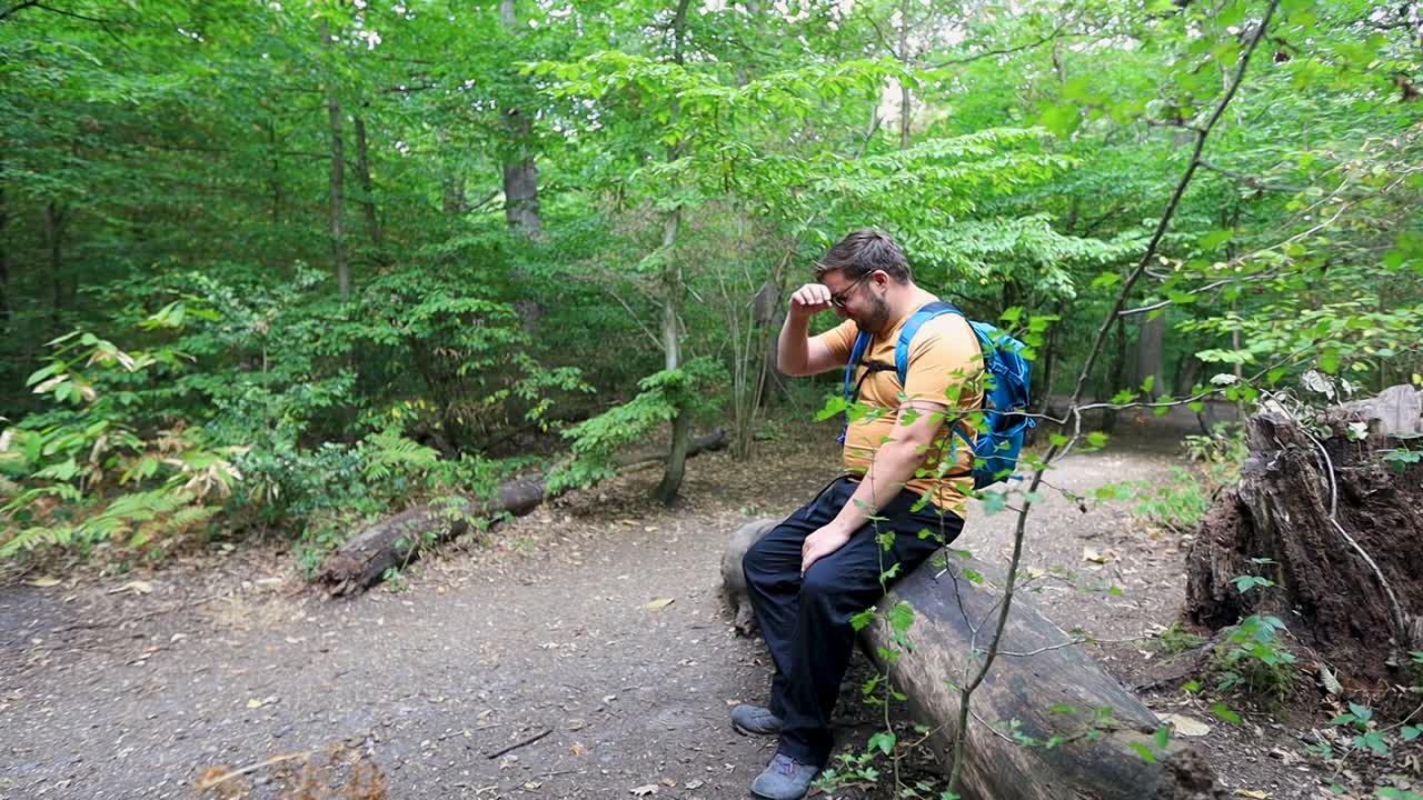 Tired traveler sits down on fallen log surrounded by dense forest, side slow motion view