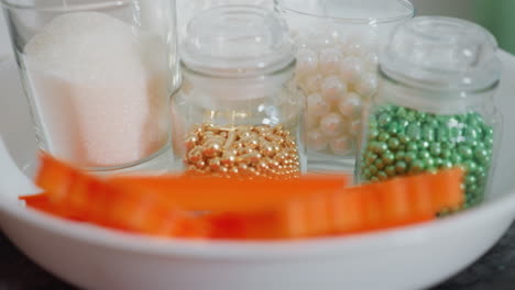 close-up of vibrant edible beads in gold, green, and pearl colors stored in glass jars, sugar in glass cup, orange cookie cutters, and baking tools arranged in white bowl on kitchen countertop