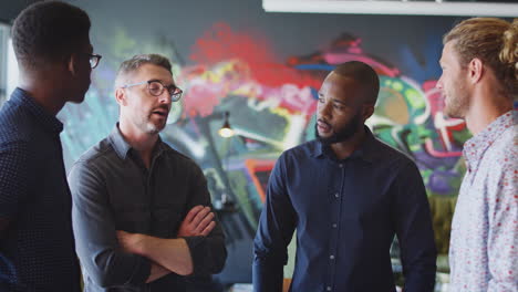 four male creatives stand talking in front of a mural in their workplace, waist up