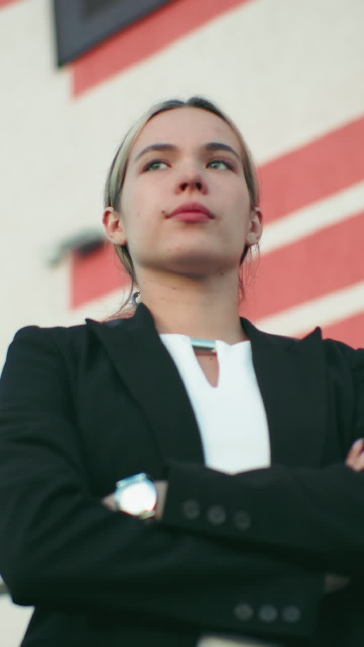 Young real estate manager in black blazer folds hands standing confidently in front of organization with red and white striped wall, security camera on building, leadership, and success