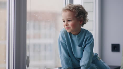 chubby slavic little boy with charming curly hair is sitting on windowsill in room in snowy day looking out window