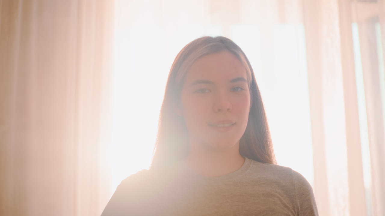 Lady in ash top stands calmly with soft sunlight glowing behind sheer curtains, creating gentle backlight effect highlighting natural face expression with serene indoor atmosphere