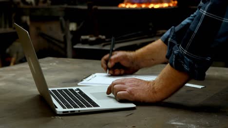 blacksmith writing on clipboard while using laptop in workshop 4k