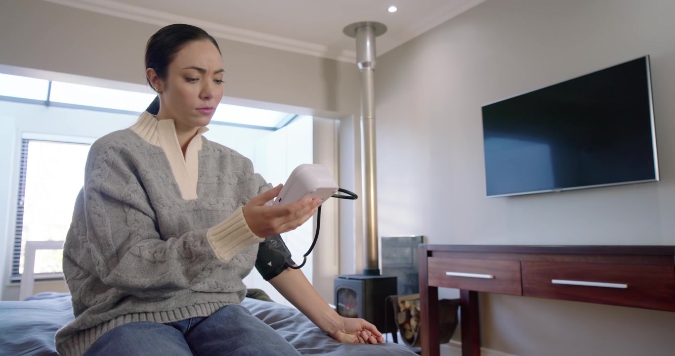 Asian woman sitting at home, wearing cuff on arm, reaching for monitor, checking blood pressure