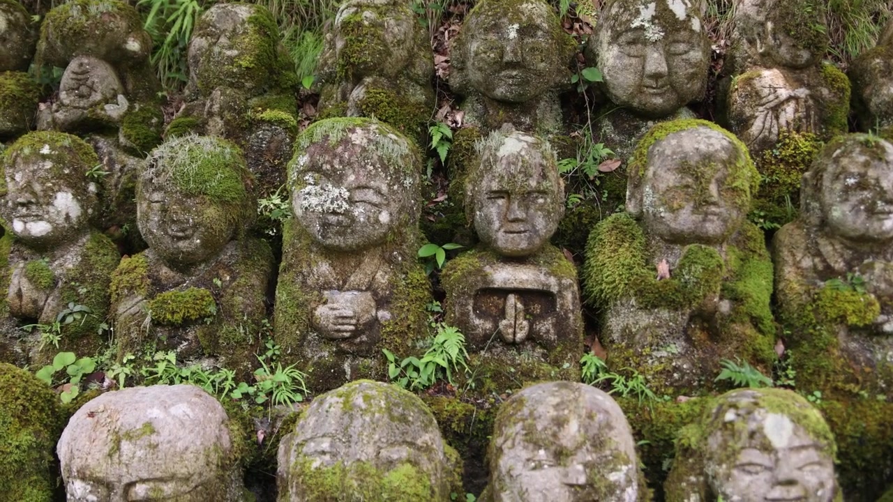 Vista ravvicinata di numerose figure buddiste in pietra consumate dalle intemperie, coperte di muschio e circondate da vegetazione, che creano una scena religiosa contemplativa nella storica Kyoto, Giappone.