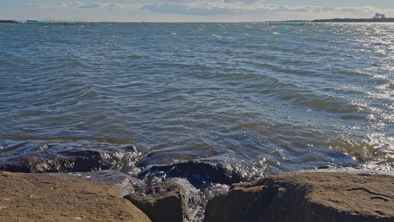 Medium shot of choppy, dark blue water breaking against large, rugged rocks near the edge of the bay