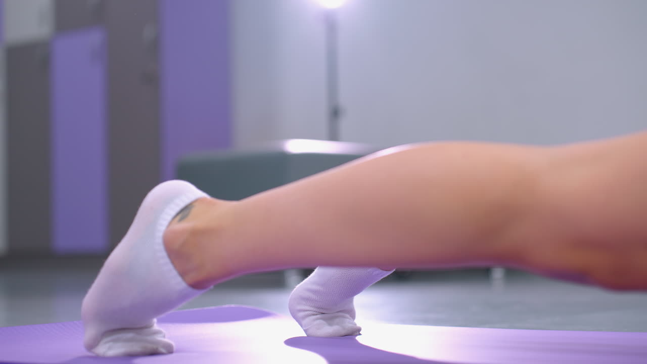 색 양말을 입고 보라색 매트 위에서 스트레칭 운동을 하는 다리의 클로즈업, 링 라이트가 체육관 공간을 비추고, 캐스팅 반사, 배경에는 다채로운 캐비이 있습니다.