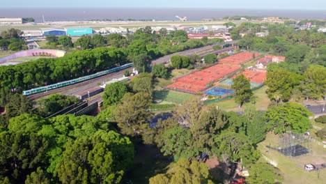 Vista-Aérea-De-Las-Canchas-De-Tenis-De-Tierra-Batida-Y-El-Tren-De-Transporte-En-Movimiento-En-Buenos-Aires,-Argentina