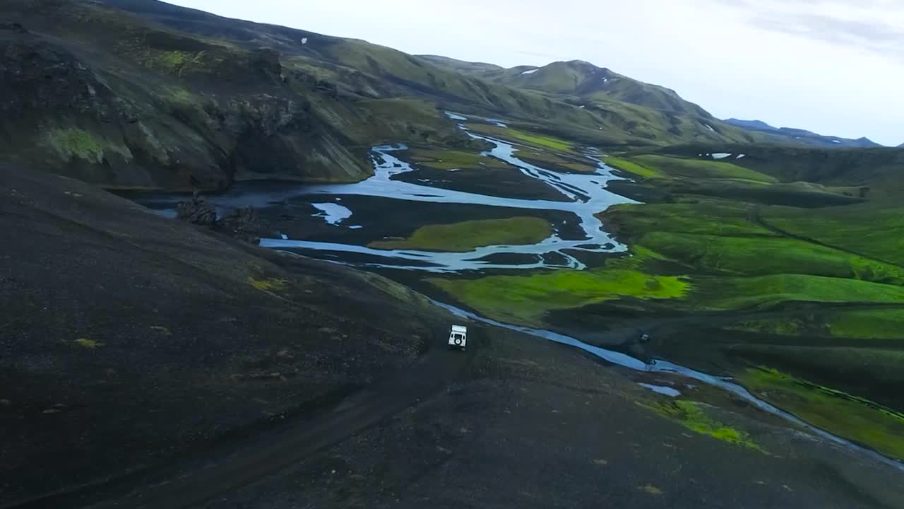 Aerial drone flying over and besides a white 4x4 truck Jeep vehicle driving in mountains with fresh water rivers around in Greenland or Iceland during a cloudy day. Moss and green grass growing around