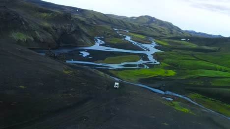 aerial drone flying over and besides a white 4x4 truck jeep vehicle driving in mountains with fresh water rivers around in greenland or iceland during a cloudy day. moss and green grass growing around