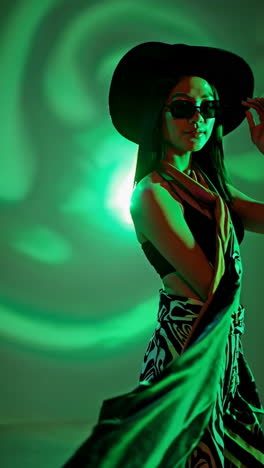 stylish woman in hat and sunglasses posing under green light