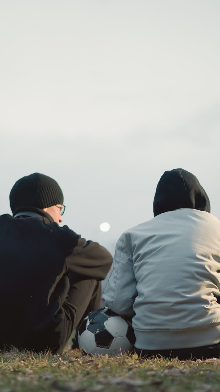 Two boys are seated on a grassy field, one wearing a gray jacket resting his left hand on a soccer ball, with blur light view and building in the background