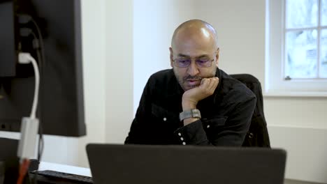pensive south asian office worker resting chin in hand while gazing intently at laptop screen, lost in thought amidst a busy workspace