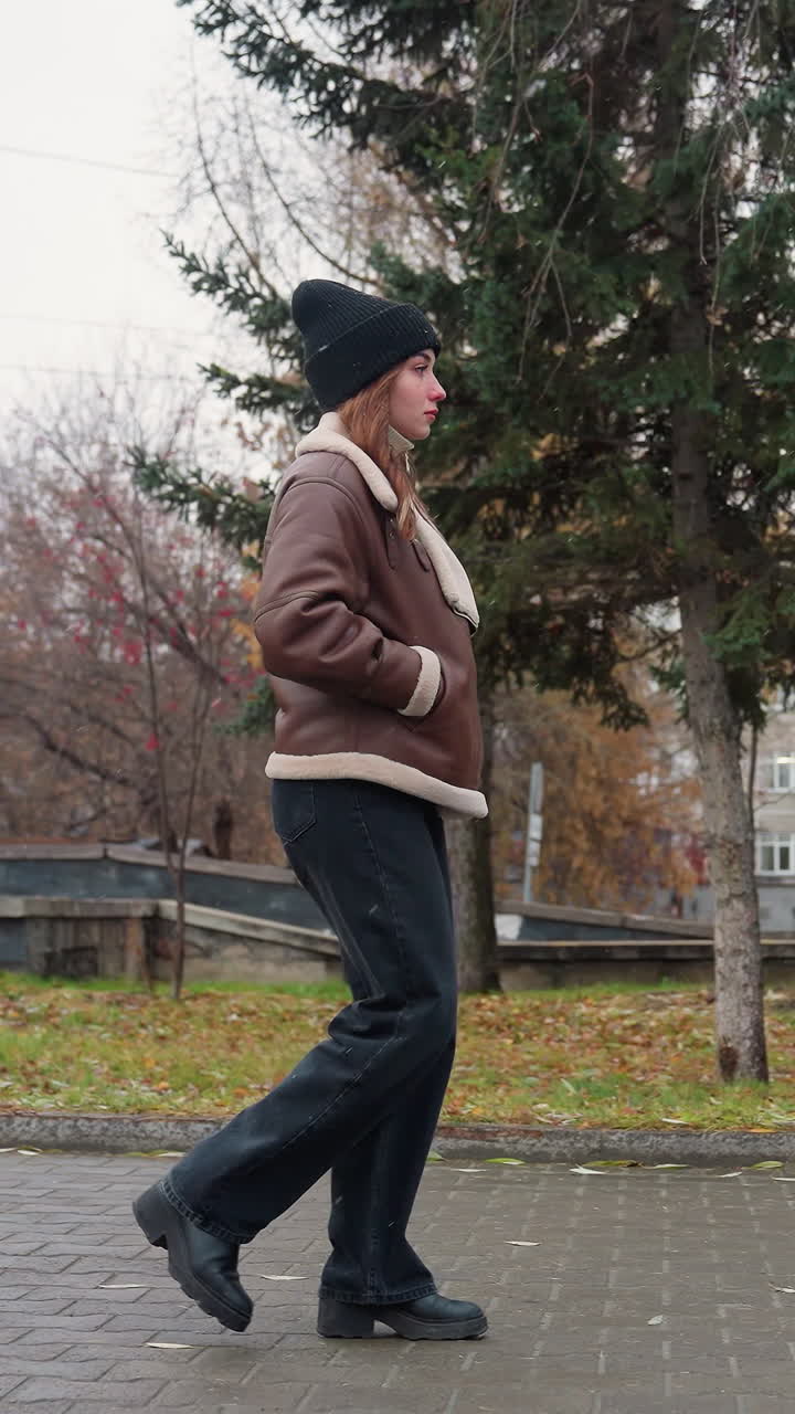 Side view of student in black knit cap, brown shearling jacket, and black trouser walking calmly through paved urban park on cold overcast day with light snowfall and colorful autumn background