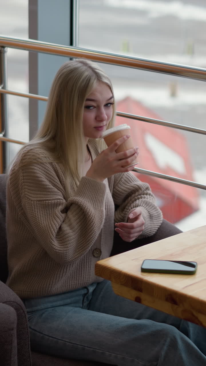 Elegant kvinde nipper til kaffe fra en kop, mens hun kigger ud gennem vinduet. Telefon hviler på et træbord i en moderne indkøbscenter-café med bymiljø i baggrunden, blød naturlig belysning og rolig stemning.