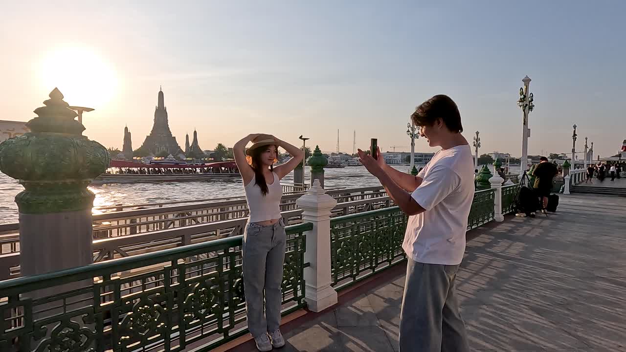 Un uomo fotografa la sua ragazza mentre posa sul lungofiume al tramonto, con una calda retroilluminazione e un'atmosfera serena