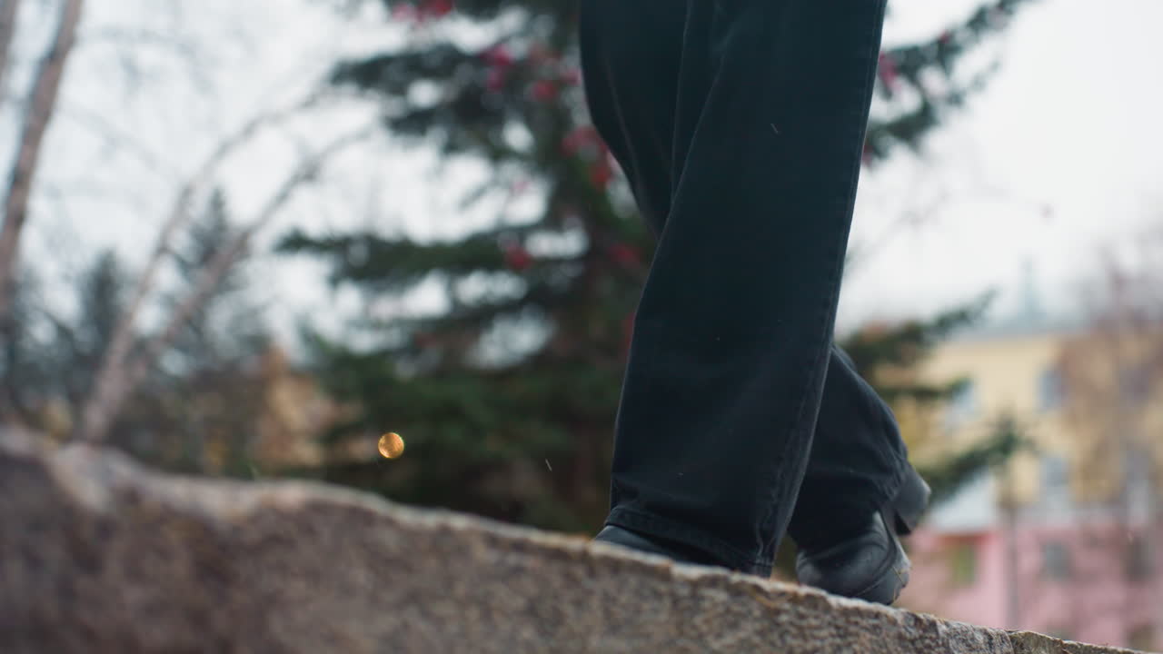 Person in black trousers and black boots carefully walking on wide wet stone path during rainy autumn day, maintaining balance amidst rain and falling leaves, enjoying nature