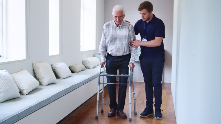 Male nurse helps senior man using walking frame, full length