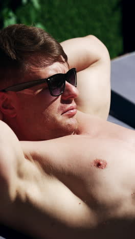 content topless caucasian man wearing sunglasses lies on the lounge. male holding hands behind his head and rises head to look around. vertical video
