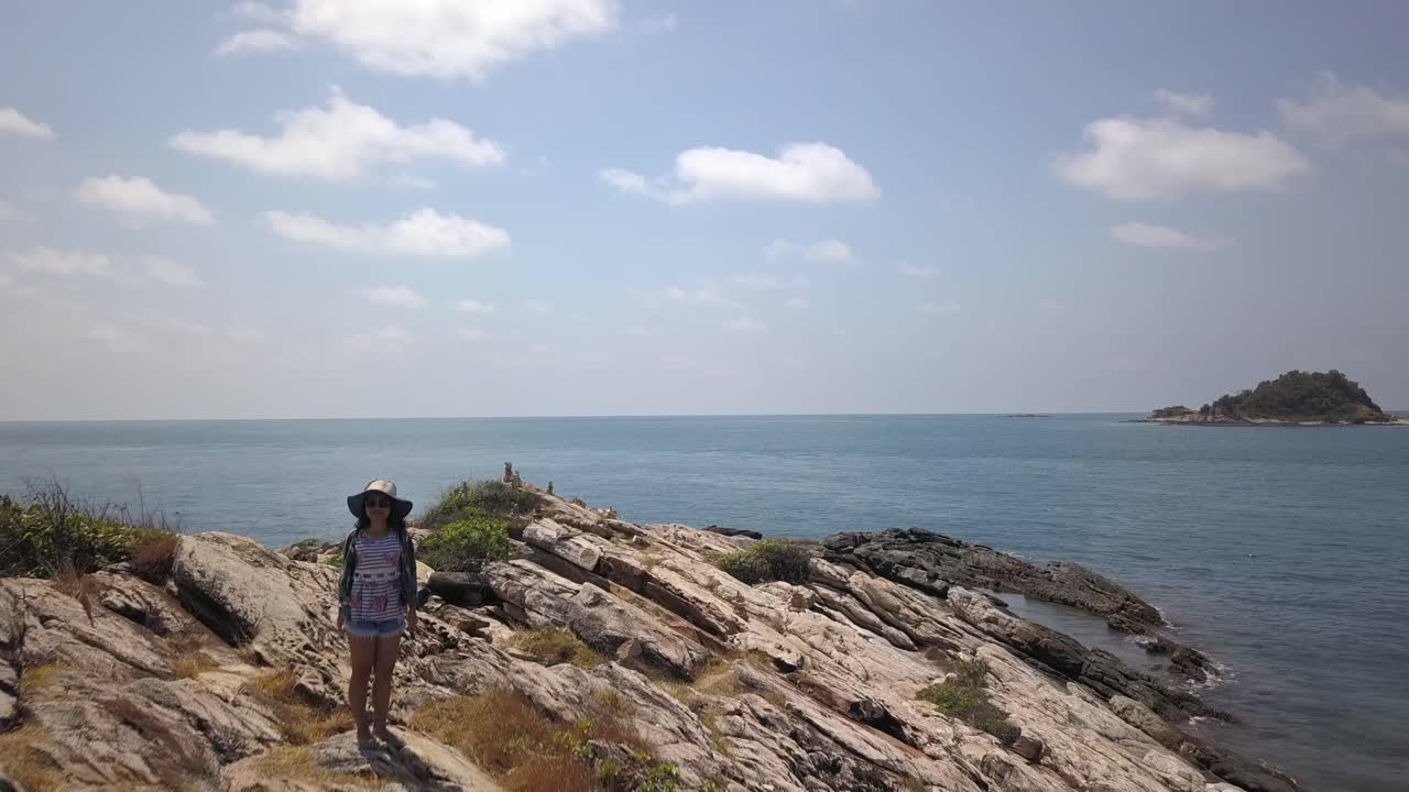 공중: 태국의 바위가 많은 관점에 서 있는 젊은 태국 여성을 드러내며 고도를 약간 낮추는 고고도 돌리 아웃 샷, 카메라는 지면에 가까워지고 멀어집니다.