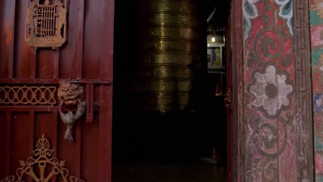 a golden tibetan prayer wheel inside a temple by the doorway with people walking passed inside - mid shot