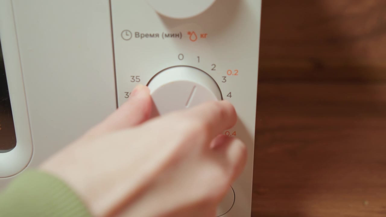 Primo piano che mostra una mano che ruota la manopola di controllo inferiore sul pannello di un microonde bianco, con la sfocatura della punta del dito che cattura la regolazione attiva, mentre le marcature numerate rimangono chiare accanto a una calda luce da cucina.