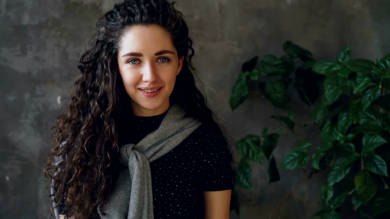 Close-up portrait of yougn attractive girl looking into camera and smiling while standing against grey wall and large green plant