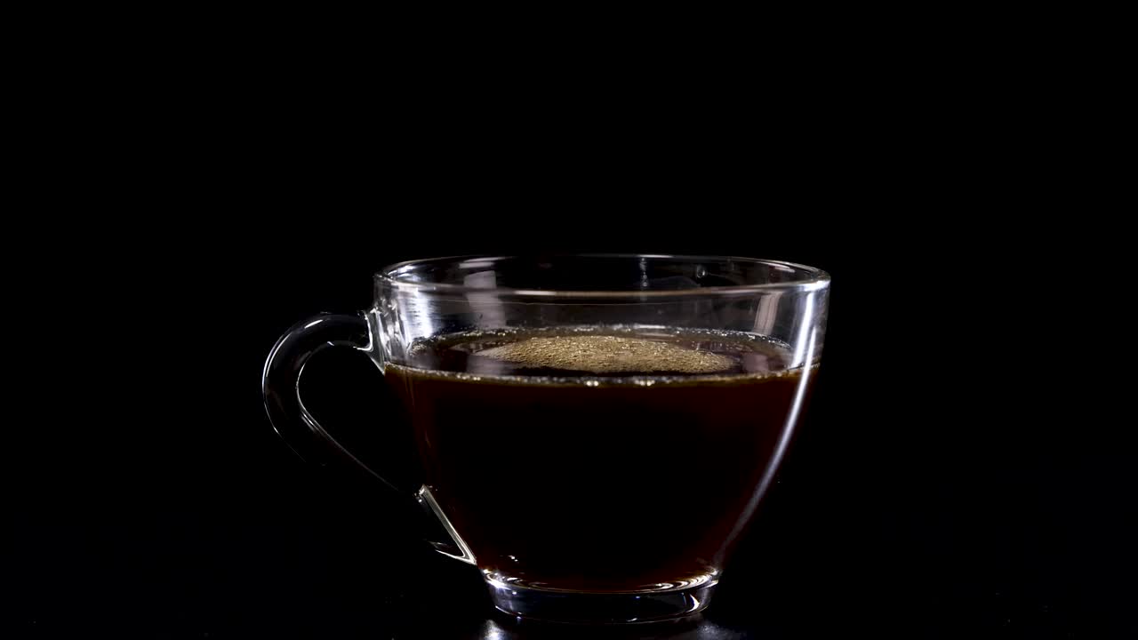 Coffee is poured into a glass cup against a black background, highlighting the rich color and texture of the liquid