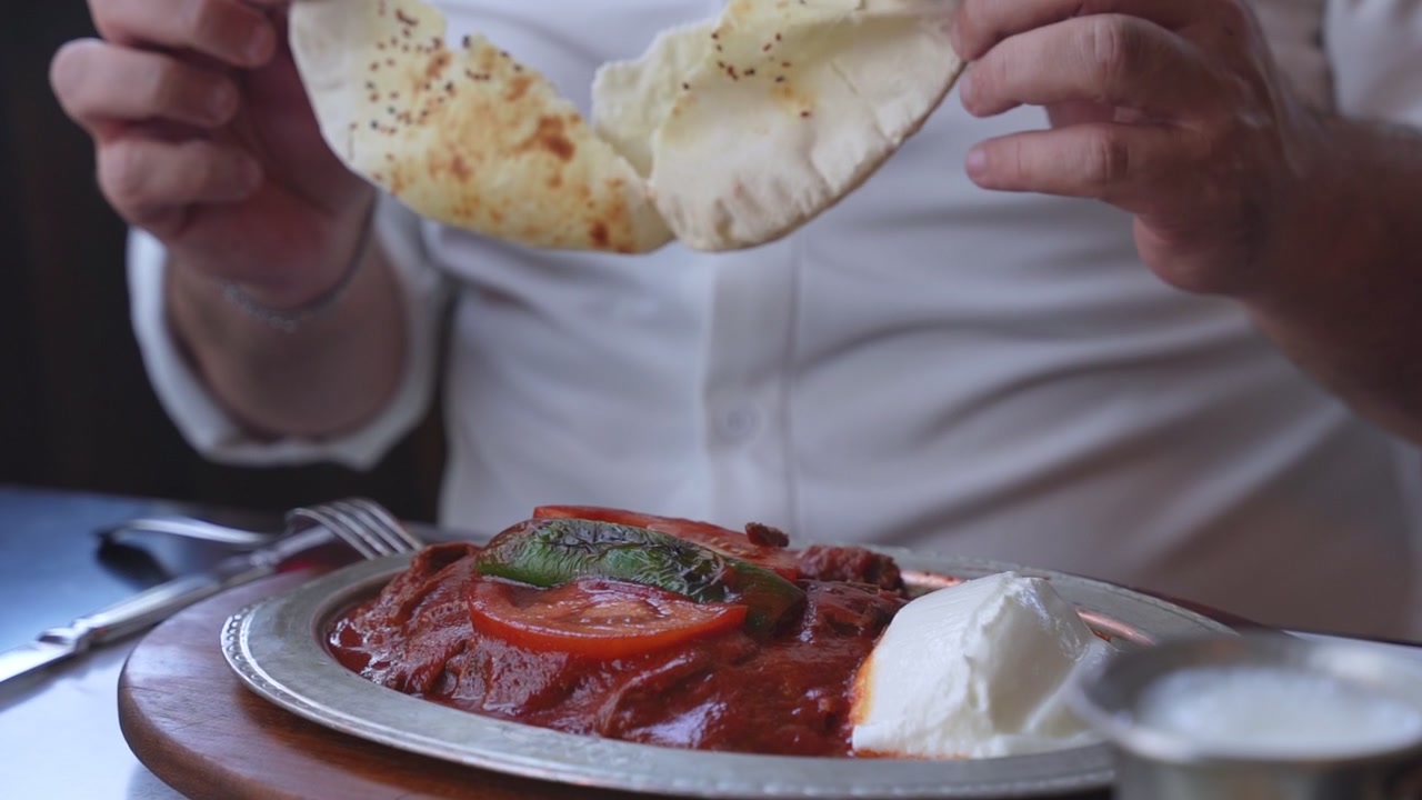 Close up shot of a man in a white shirt tearing fresh flatbread over a traditional Turkish Iskender kebab plate in a restaurant. The dish features meat, tomato sauce, and yogurt under natural light.