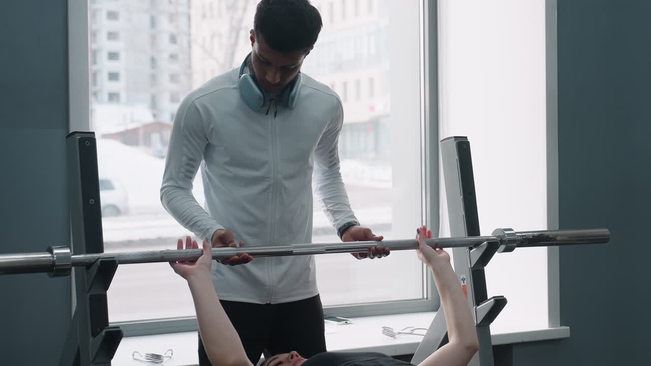 young woman lies on bench holding barbell while male trainer stands nearby offering support inside modern gym with large windows, natural daylight, and passing car visible through glass