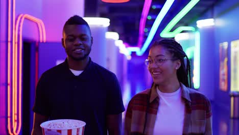 happy young african american woman and man walking by neon hall with bucket of popcorn, chatting at cinema