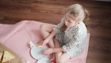 overhead view of little kid sitting cross-legged on pink bedspread eating slice of pizza, holding plate with crumbs in cozy indoor setting, casual mealtime scene with natural lighting