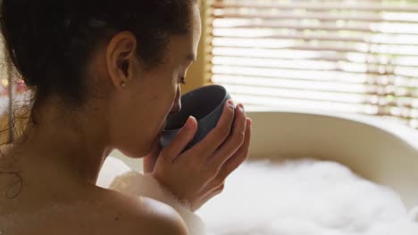 close up of relaxed biracial woman with vitiligo sitting in bath with foam and drinking coffee