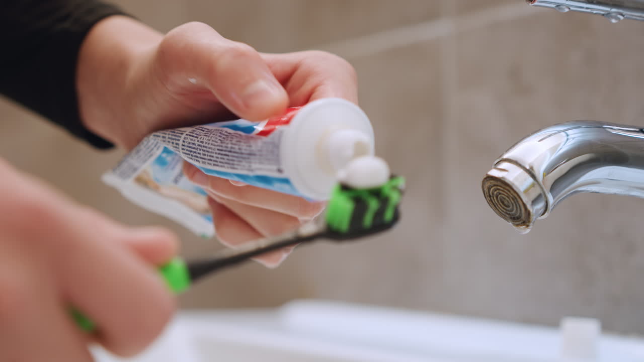 Close up hands applying toothpaste to green toothbrush near chrome faucet over porcelain sink, bathroom tile background, steady squeeze from tube, white paste on bristles, morning dental routine, calm