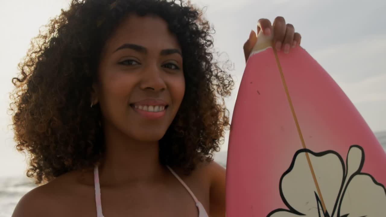 Front view of African American female surfer standing with surfboard on the beach 4k