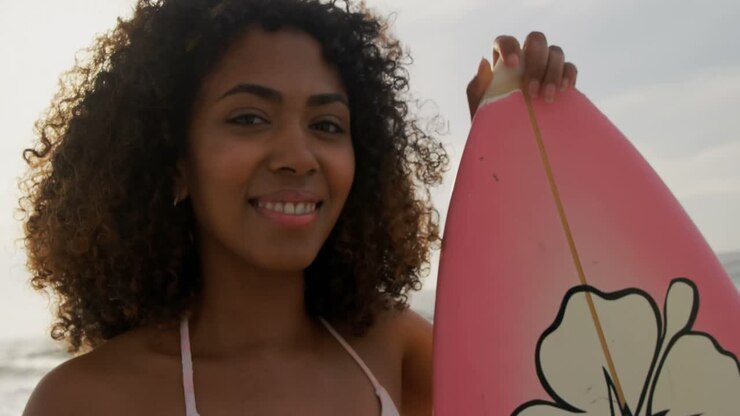 Vista frontale di surfista afroamericana in piedi con la tavola da surf sulla spiaggia 4K