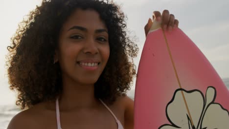 vista frontal de una surfista afroamericana de pie con una tabla de surf en la playa 4k